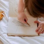 Small Business Owners - From above of small girl in dress drawing with pencil in notebook while mother using laptop on bed near
