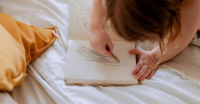 Small Business Owners - From above of small girl in dress drawing with pencil in notebook while mother using laptop on bed near
